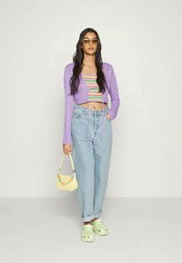 Casaco cropped tricotado roxo, top cropped às riscas, jeans de cintura alta azul clara, tamancos verde pastel e uma pequena mala amarela com detalhes coloridos.