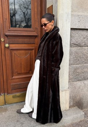 Moteris dėvi tamsų kailinį paltą, akinius nuo saulės, tamsią palaidinę ir plačias baltas kelnes, stovinti šalia medinių durų su akmenine siena fone.