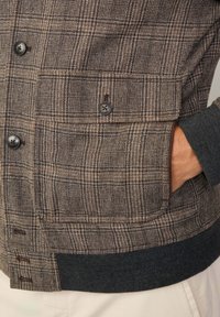 Chaqueta de cuadros marrón con una tela texturizada, acentos de botones negros y un dobladillo de canalé oscuro. Incluye bolsillos laterales y un cierre de botón.