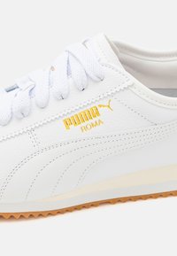 Baskets blanches Puma Roma avec logo doré, lacets blancs, dessus en cuir texturé et semelle en caoutchouc marron clair.