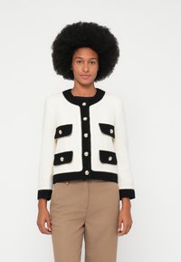 Veste tricotée en crème et noir, avec un col rond, des boutons décoratifs et quatre poches avant avec un passepoil contrastant.