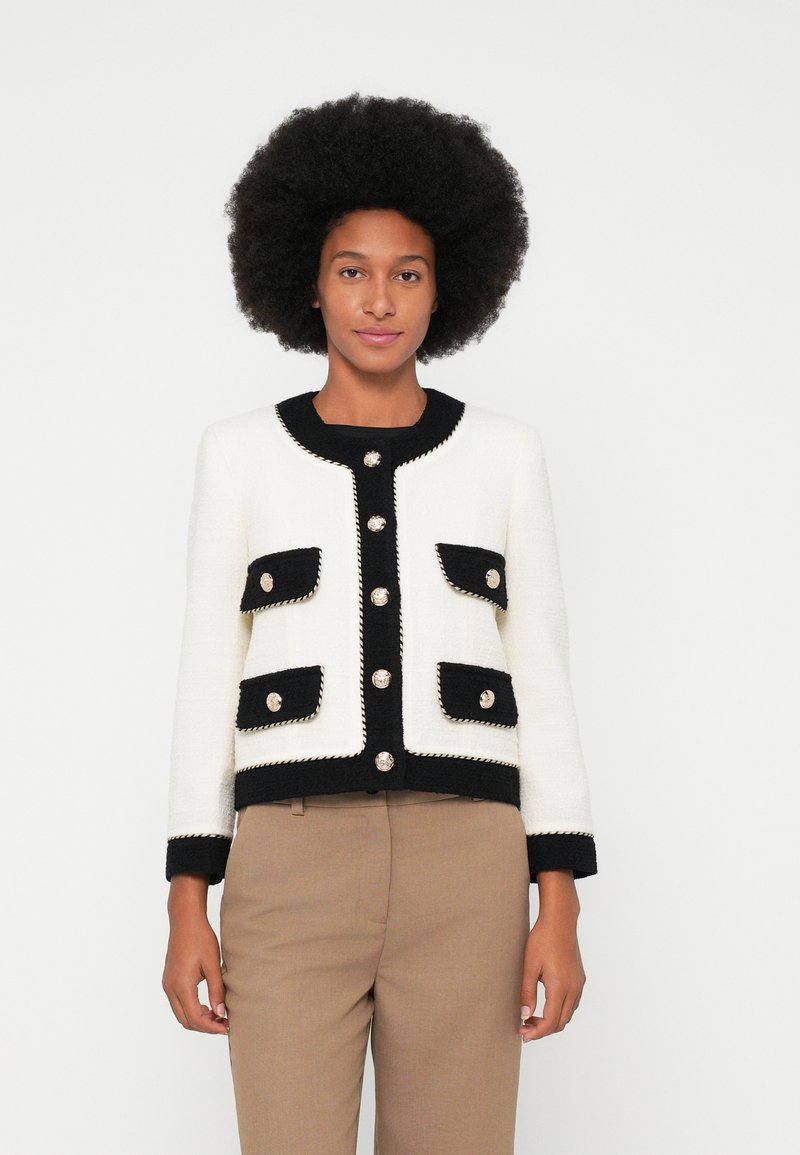 Veste tricotée en crème et noir, avec un col rond, des boutons décoratifs et quatre poches avant avec un passepoil contrastant.
