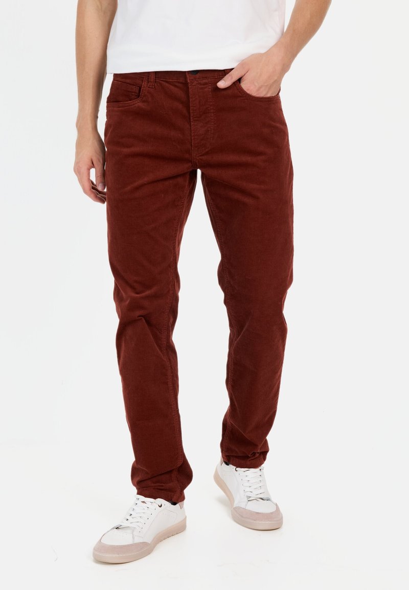 Burgunderfarbene Cordhosen im Slim-Fit mit fünf Taschen und einem Knopfverschluss. Getragen mit weißen Sneakers mit einem hellrosa Akzent.