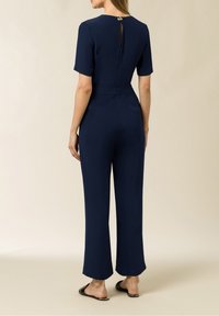 Tuta navy con maniche corte, vita aderente, gambe larghe e dettaglio a chiave sul retro. Realizzata in un tessuto liscio, con hardware minimale.