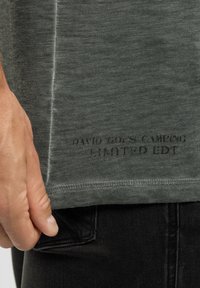 Close-up of a hand holding black denim jeans pocket edge, wearing a green shirt with "David Cross Camping Limited Edt" printed near the hem.