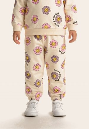 Kind draagt een crèmekleurige trui en broek met lachende bloemmotieven en de tekst "have a nice day", gecombineerd met witte sneakers.