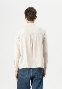 Beige long-sleeve button-up shirt featuring a textured fabric, back pleat, and a standard collar, paired with blue denim jeans.