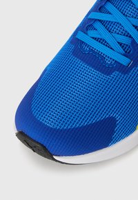 Gros plan sur une chaussure de sport en maille bleue avec des lacets bleus et une semelle noire et blanche sur fond blanc.