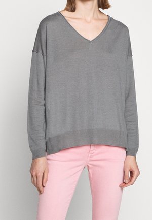 Kvinde iført en løs grå V-hals trøje og tætsiddende lyserøde bukser, stående mod en ensfarvet lys baggrund.