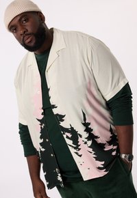 Korte mouw knoopshirt in crème met een bosontwerp in zwart en roze. Draagt over een donkergroene lange mouw shirt. Zachte stof, losse pasvorm.