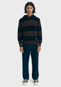 Personne avec des cheveux bouclés portant un sweat à capuche tricoté rayé marron et noir, un jean bleu foncé et des baskets noires et blanches, se tenant contre un fond blanc.
