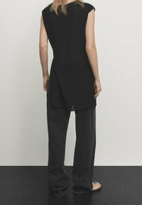 Vrouw die blootsvoets met haar rug naar ons gekeerd staat, in een mouwloos zwart top met zijsplitten en losse zwarte broek tegen een effen achtergrond.