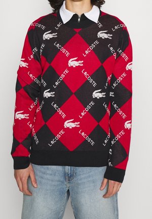 Osoba nosząca czerwono-czarny sweter w argyle z powtarzającymi się białymi logo Lacoste i motywami krokodyla, w połączeniu z jasnoniebieskimi dżinsami.