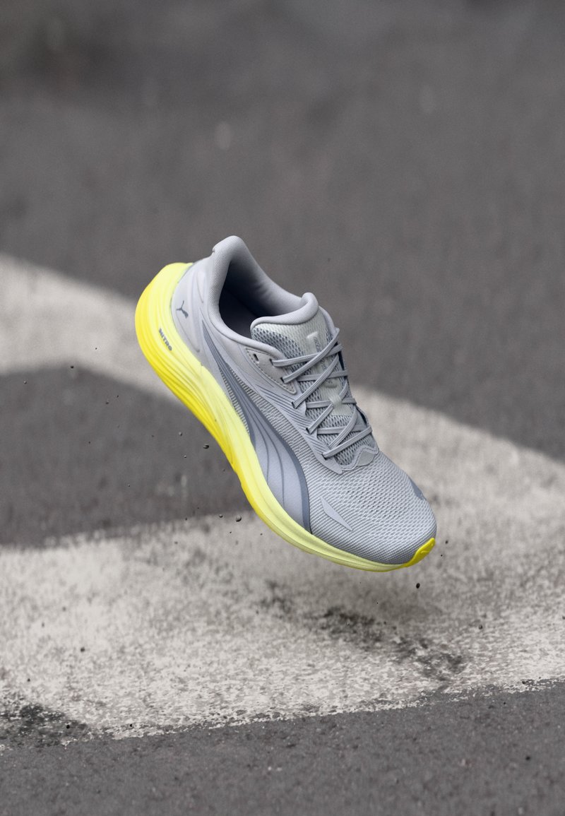 Chaussure de course grise et jaune en l'air au-dessus d'une surface en asphalte marquée de lignes blanches de route.