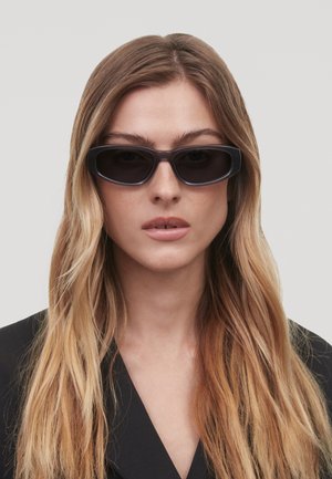 Lunettes de soleil noires rectangulaires avec une finition brillante, dotées de verres foncés et de grosses montures, portées par une personne en blazer noir.
