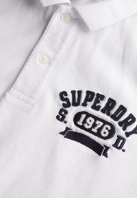 Camisa polo blanca con una tela texturizada, que presenta un logotipo bordado en azul marino oscuro que dice "SUPERDRY 1976" y un cuello abotonado.
