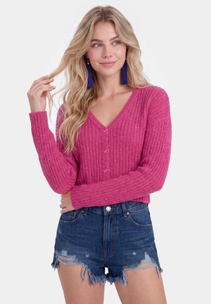 Blonde vrouw draagt een felroze gebreid vest met knopen en blauw versleten denimshorts, poseert met één hand in haar haar en een lichte glimlach.