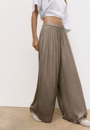 Personne portant un pantalon ample taupe à jambes larges, un top court blanc et des chaussures blanches tissées slip-on, debout devant un fond uni.