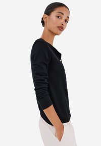 Černý cardigan vyrobený z měkké tkaniny, s kulatým výstřihem, zapínáním na knoflíky a dlouhými rukávy s ribbovanými manžetami.