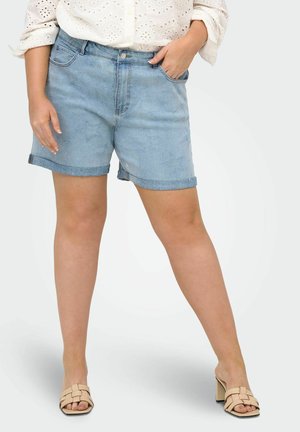 Kvinde iført lyseblå denimshorts med stjernemønstre, en hvid bluse med hulmønster og beige sandaler med blokhæl, står med den ene hånd i lommen.