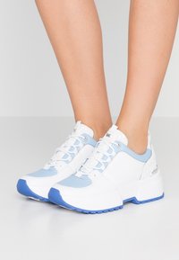 Vita och ljusblå sneakers med en texturerad bakpanel, kraftig sula och kontrasterande blå gummidetaljer. Snörningsdesign med metalöglor.