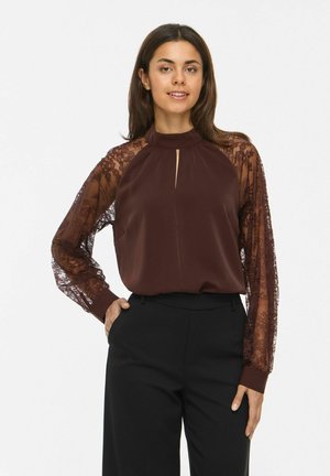 Une femme porte un chemisier bordeaux à manches en dentelle transparente et un pantalon noir sur un fond blanc uni.