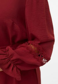 Haut bordeaux en tissu texturé, avec des manches froncées et un délicat bord en dentelle aux poignets. Détails soignés et tombé doux.