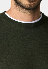 Tmavě zelený pletený svetr s kulatým výstřihem. Má texturovanou tkaninu a kontrastní bílý vnitřní límec. Jemný povrch.