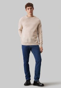 Maglione in maglia beige chiaro con scollo rotondo e orlo a costine, abbinato a jeans blu scuro e scarpe nere con lacci.