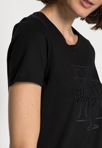 Kvinde iført en sort kortærmet t-shirt med et tekstureret "Hilfiger" logo på brystet, beskåret under hagen og over taljen.