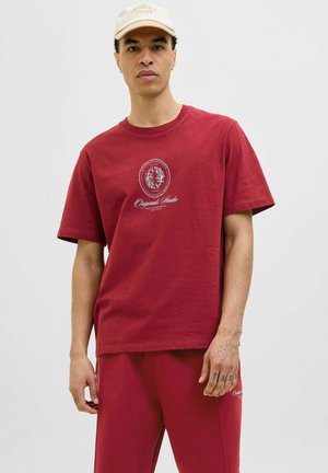 Tricou roșu din bumbac, cu gât rotund, mâneci scurte și un logo grafic central. Textură netedă cu ușoară elasticitate. Pantaloni roșii asortați.