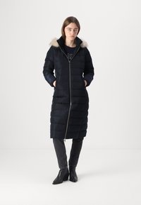 Lungo cappotto puffo blu navy con zip frontale e cappuccio con pelo, caratterizzato da un design trapuntato e tasche laterali. Indossato sopra una maglietta nera e jeans.