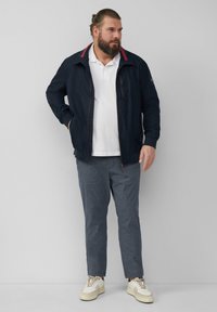 Marineblauwe jas met rode kraagaccenten, ritssluiting en twee zijzakken. Witte polo eronder, gecombineerd met grijs geruite broek en sneakers.