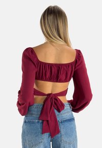 Bordeauxfarbenes Cropped Top mit Puffärmeln, geraffter Textur und gebundenem Rücken. Kombiniert mit hellblauen High-Waist-Jeans.