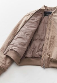Giubbotto bomber beige con una superficie esterna liscia, fodera interna trapuntata, polsini a coste e zip frontale. L'interno presenta una texture morbida.