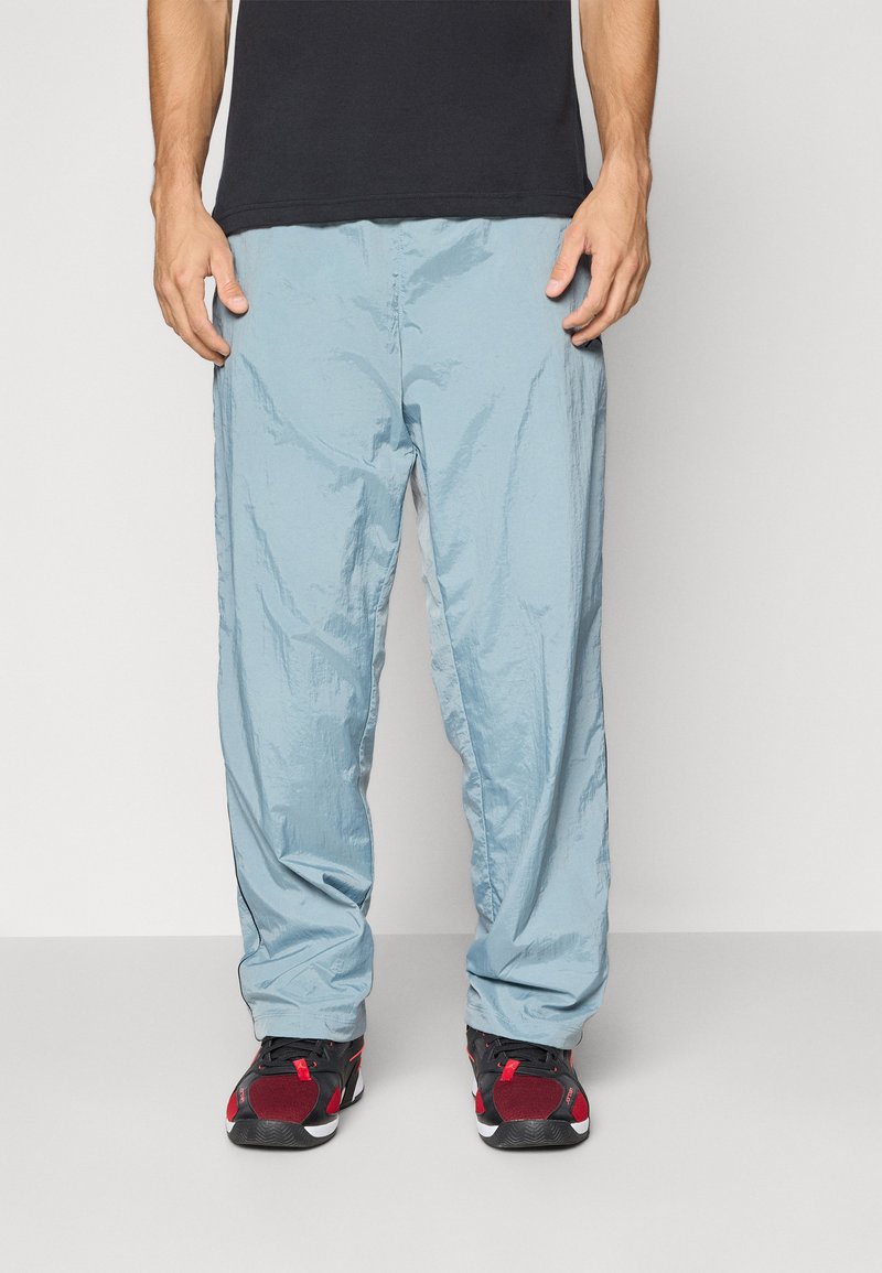 Mand iført lyseblå løse bukser, sorte og røde sneakers samt sort t-shirt, stående foran en ensfarvet grå baggrund.