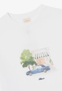 White cotton t-shirt featuring a watercolor illustration of a blue car, a tree, and a storefront with a striped awning, labeled "Blkvis."