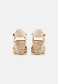 Sandalias de cuña con parte superior de yute beige y tacón texturizado en yute. Presentan una correa ajustable en el tobillo y un diseño de parte trasera abierta.