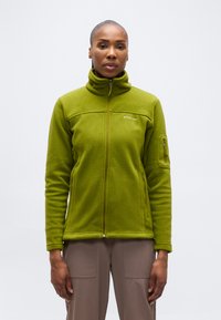 Γυναίκα που φοράει πράσινο μπουφάν φλις με φερμουάρ, ψηλό γιακά και τσέπες με φερμουάρ, στέκεται μπροστά σε απλό φόντο.