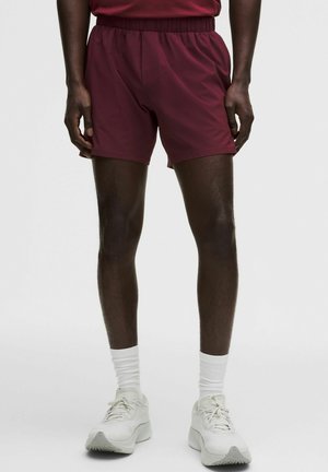 Bordeauxrote Sportshorts mit elastischem Bund und leichtem Stoff, kombiniert mit weißen Socken und hellen Turnschuhen.