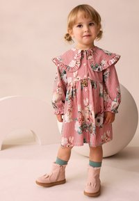 Rot-weißer gestreifter Kleid mit floralen Mustern und einem Rüschkragen, kombiniert mit rosa Knöchelboots und grünen Socken.