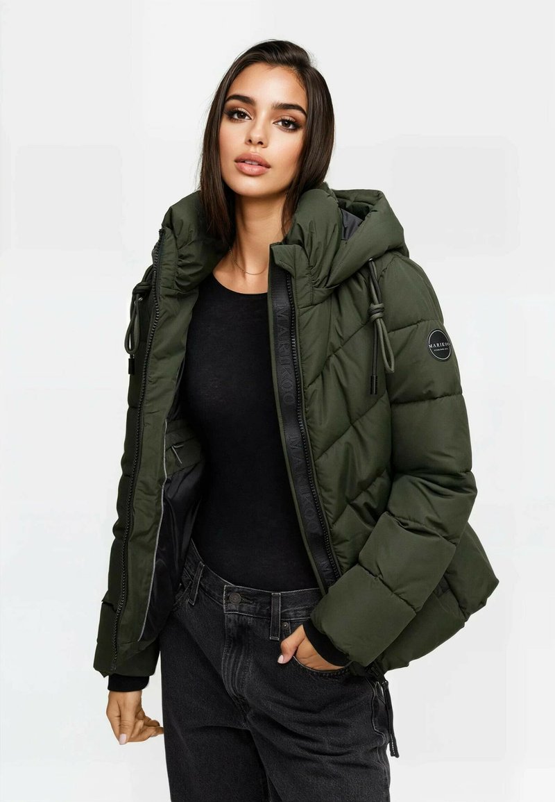 Kobieta w niezapniętej ciemnozielonej kurtce puchowej na czarnej koszulce i czarnych dżinsach, z jedną ręką w kieszeni, na jednolitym tle.