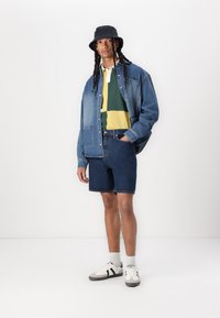 Denim jacket over a green and yellow striped polo, paired with dark denim shorts and white sneakers with black accents. Hat included.
