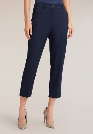 Kvinde iført marineblå cropped straight-leg bukser og beige højhælede sko stående på en neutral baggrund.