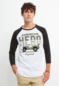 Hvid og sort raglan t-shirt med grafik af en 1944 Willys MB Jeep, tekst "AMERICAN HERO" og "Legend" samt stjerneaccenter.