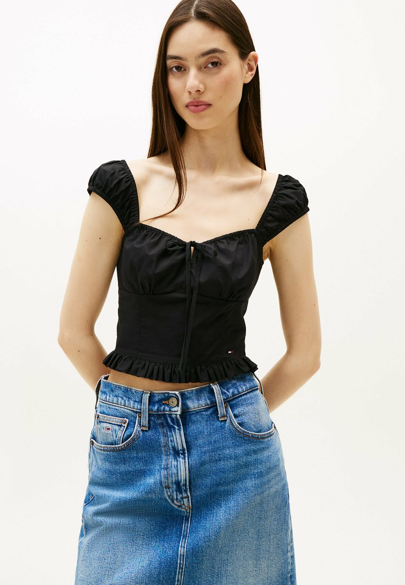 Top cropped nero con maniche corte riflettenti, scollatura quadrata e nodo centrale. Abbinato a una gonna in denim blu a vita alta con tasche anteriori.