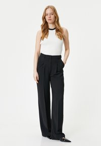 Witte geribde mouwloze top met zwarte randen, gecombineerd met hoge zwarte wijde broek. Model staat met handen in de zakken.