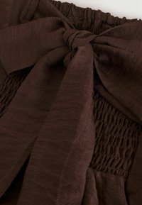 Mörkbrunt tyg som är knutet i en rosett med en smockad elastisk del, som visar mjuk textur och en subtil diagonalt vävningsmönster.