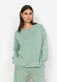 Světle zelená mikina s dlouhými rukávy, kulatým výstřihem a volným střihem. Hladká textura, mírně oversize silueta a manžety ohrnuté na zápěstích.