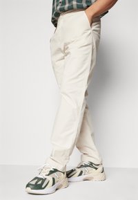 Pantalons en coton beige à coupe droite, dotés de poches latérales et d'une texture lisse, associés à des baskets de sport vertes et beiges.
